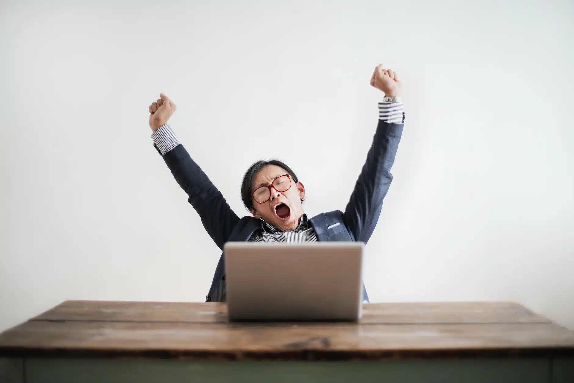 photo of yawning man with his hands up and eyes closed sitting at a table with his laptop
